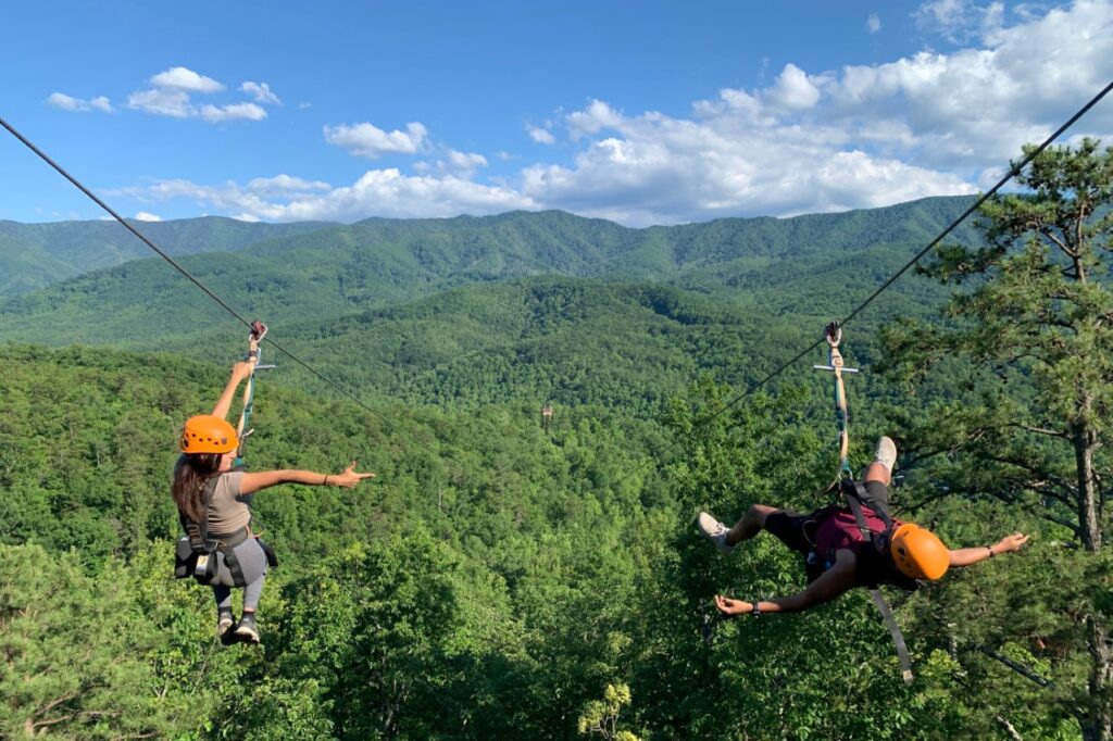 Two people riding a zipline. Woman points finger guns at man. Man plays dead.